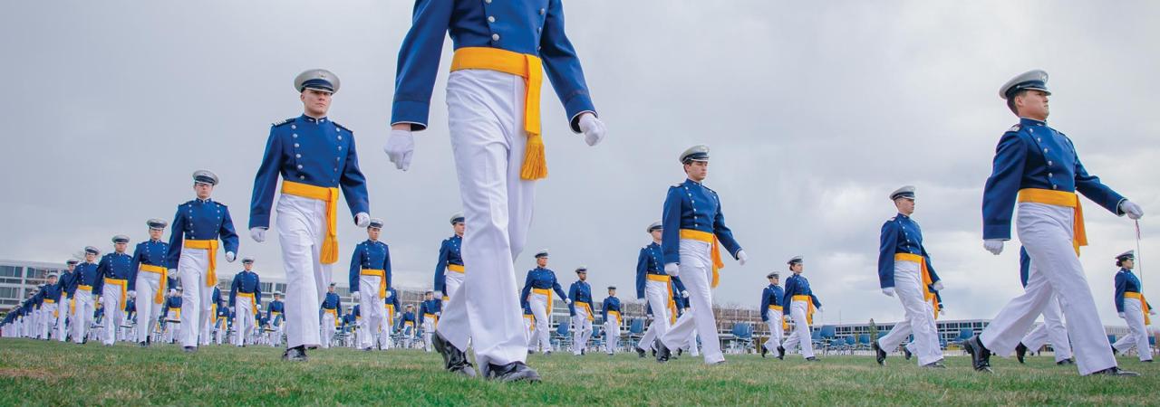 Cadets in blue and yellow uniforms, wearing yellow sashes, march on a field at graduation