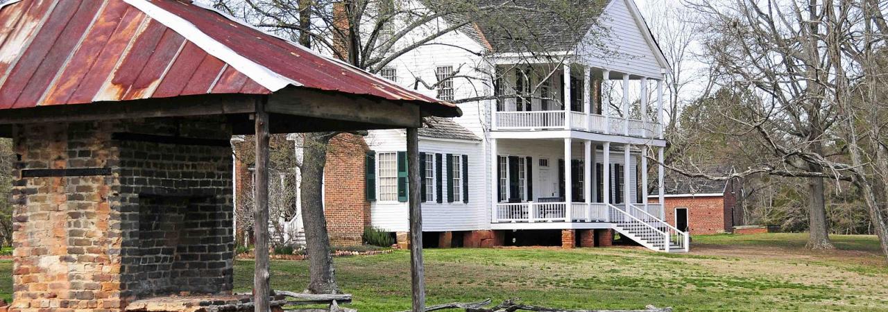 Photograph of a outdoor brick fireplace in the foreground and a two-story white building with a porch in the background.