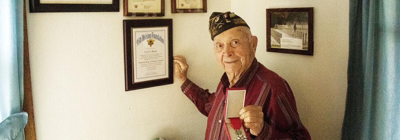 World War II veterans stands with medal in front of a wall of awards.