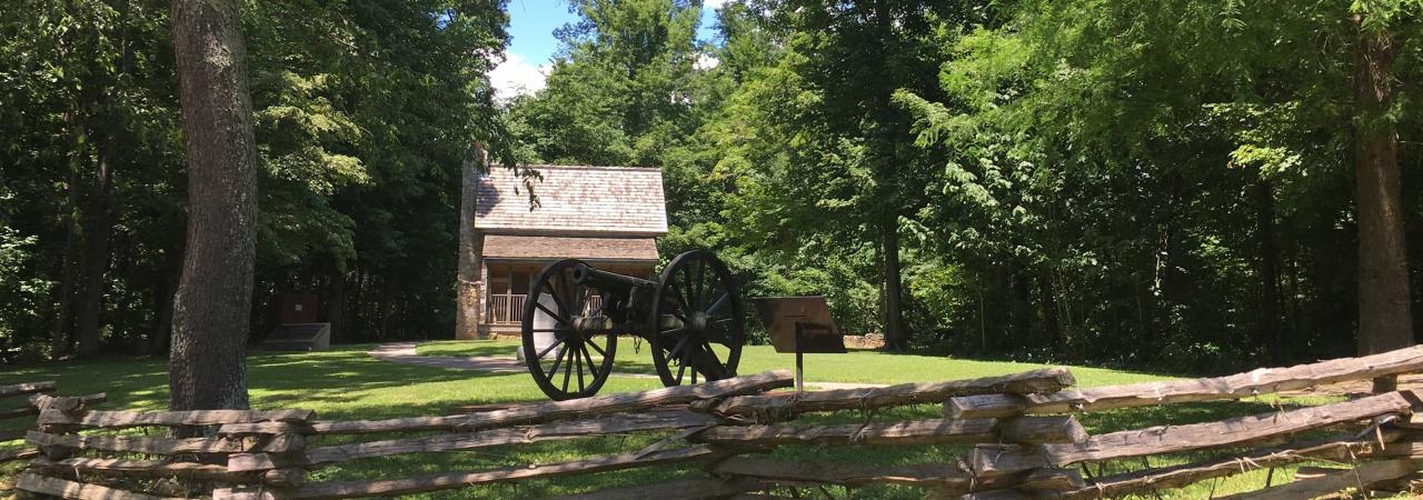 Battlefield with a cannon in the foreground and a log cabin in the background