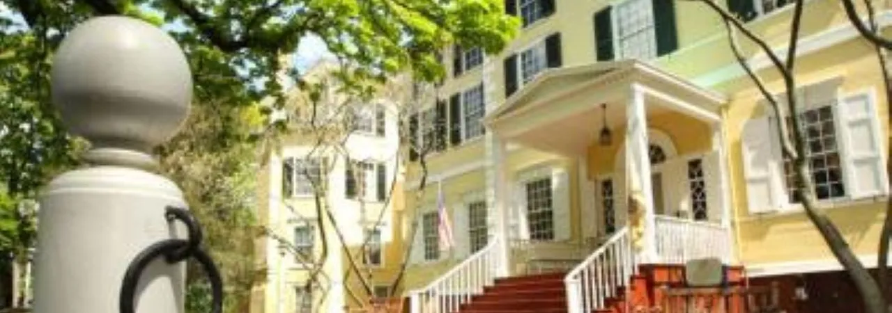 A large yellow historic house with white trim and a columned front porch stands among trees on a sunny day.