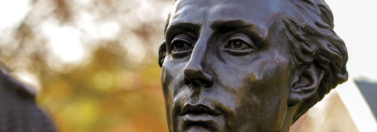 Bust of bronze statue of Revolutionary War era gentleman with foliage in the background