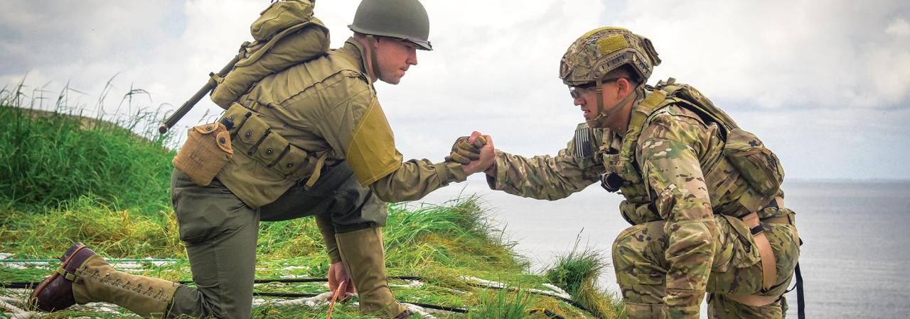 On a grassy embankment with the ocean in the background, one solider in uniform kneels and helps another up the cliff