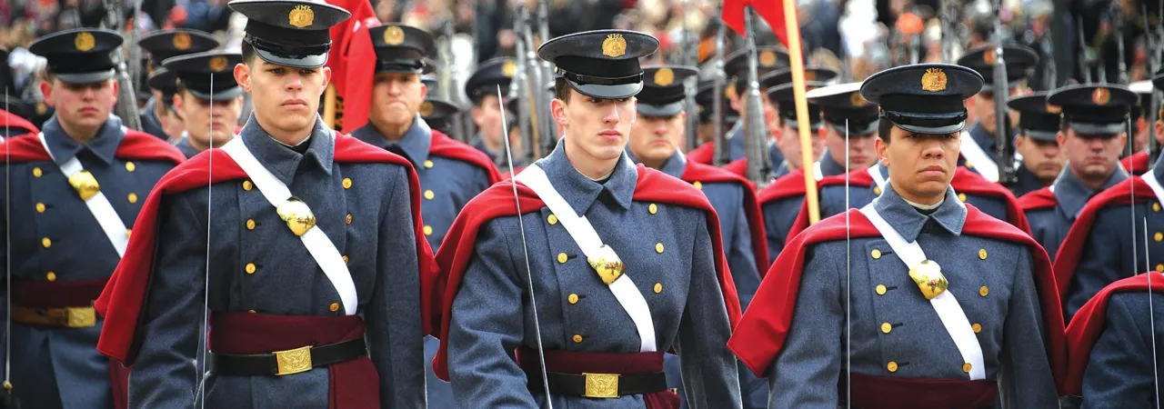 Cadets in uniform march with swords and flags with stadium audience in the background