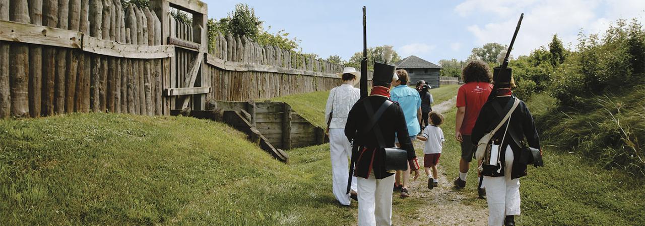 A group of tourists and two War of 1812 reenactors walk along a stone path next two a wooden fence. 