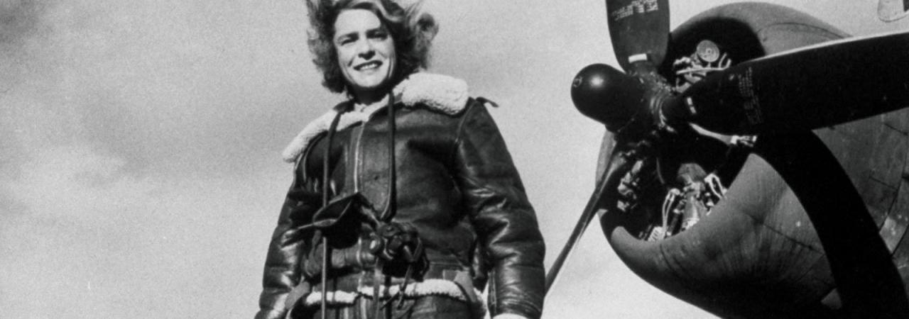 Black and white photo showing a woman in a pilot's jacket with a camera, standing in front of airplane propeller