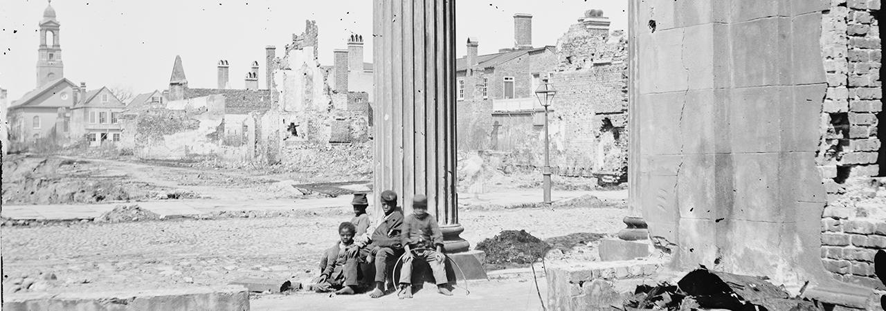 This is a black and white image of four young boys sitting barefoot and dressed in ragged clothing on the rubble of a battlefield.