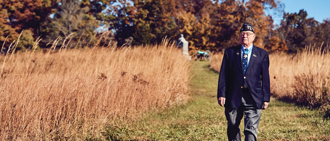 World War II Veteran in a blazer and hat walks a field with a monument in the background
