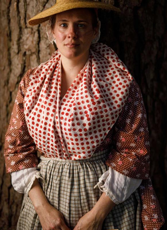 A photograph of a woman in Revolutionary War-era garb posing in front of a tree