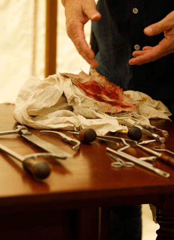 A surgeon's hands next to various Revolutionary War-era surgical tools and bloodied rags
