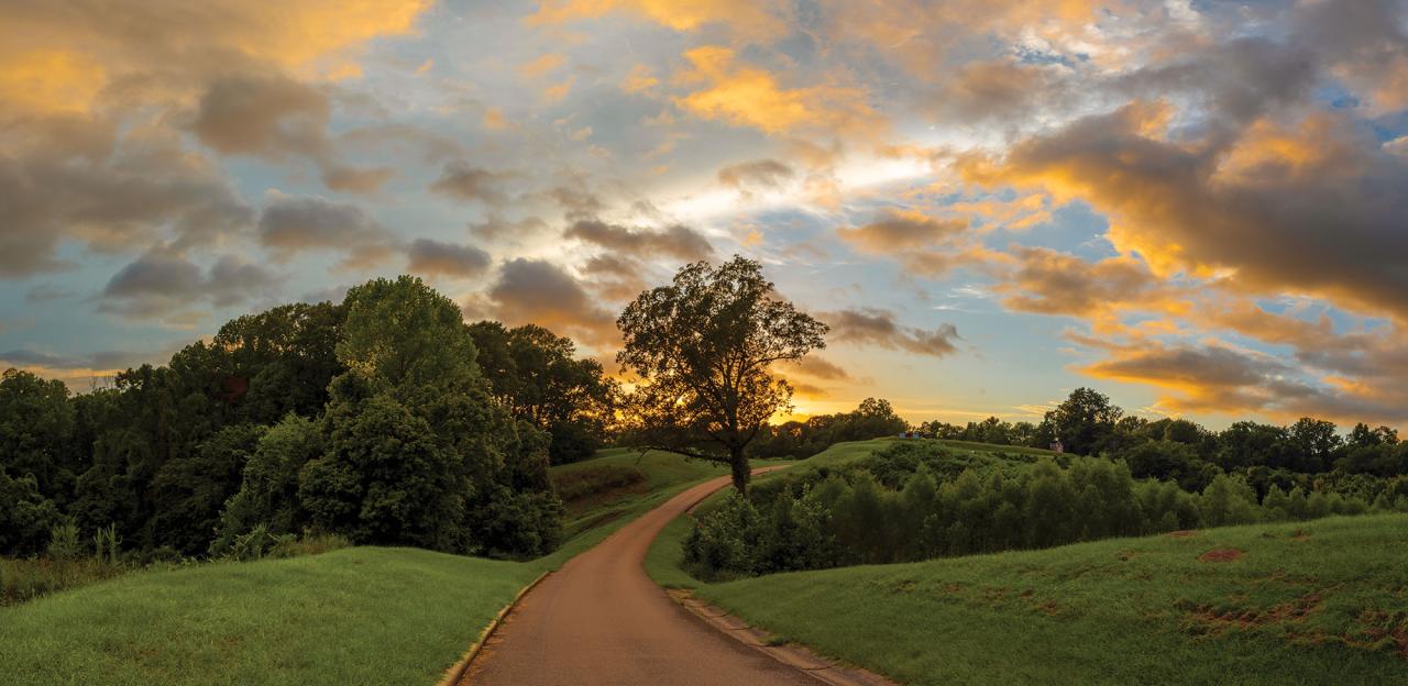 A paved path leads up a hill to a monument in the far distance, under a colorful sunset.