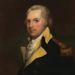 A man with white hair sits and looks at the viewer wearing the blue coat of the Continental Army with buff-colored lapels and a gold epaulet. A ruffled shirt peaks out from under his coat. 