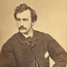 Seated portrait of John Wilkes Booth wearing a dark suit and holding a pipe in one hand.
