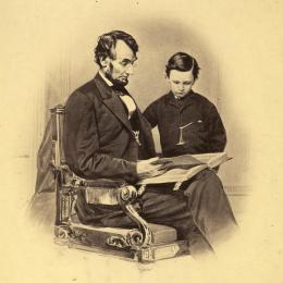 President Abraham Lincoln sits wearing spectacles beside his young son Tad, examining a book altered in the photograph to resemble a Bible.