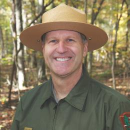 Smiling man wearing a broad-brimmed National Park Service ranger hat and green uniform, outdoors in a wooded setting.