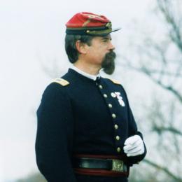 Man with dark hair and a mustache wearing a Civil War-era military uniform and red kepi-style cap.