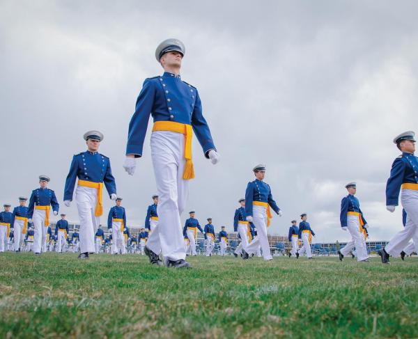 Cadets in blue and yellow uniforms, wearing yellow sashes, march on a field at graduation
