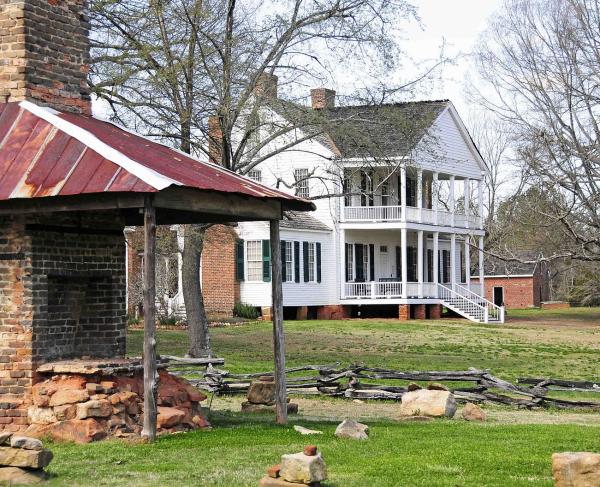 Photograph of a outdoor brick fireplace in the foreground and a two-story white building with a porch in the background.