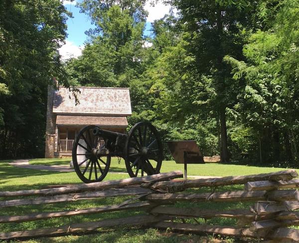 Battlefield with a cannon in the foreground and a log cabin in the background