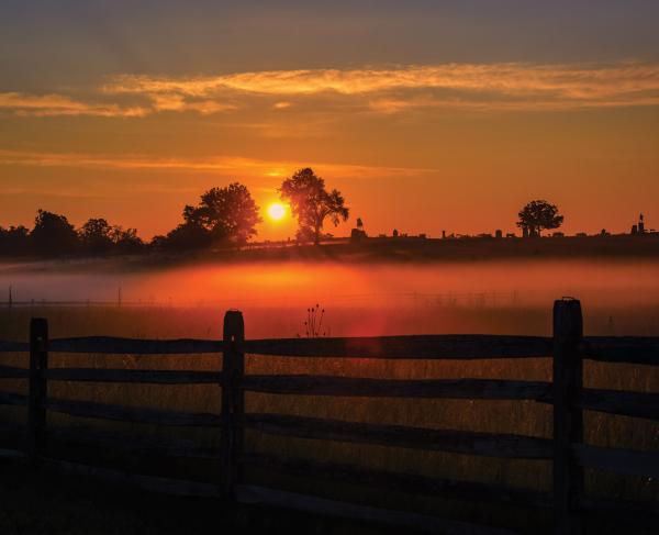 A photograph of the sun setting at Cemetery Ridge. A teeline is in the distance and a wooden fence is in the foreground.