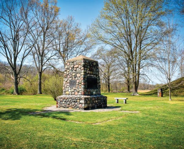 Short obelisk monument covered in rocks in a field with trees and mound seen in the distance 