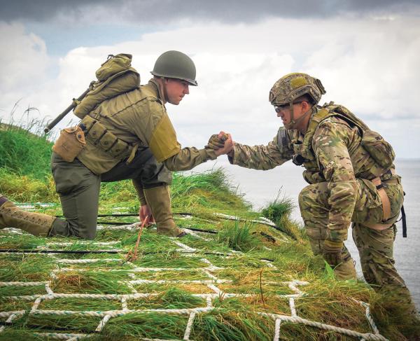 On a grassy embankment with the ocean in the background, one solider in uniform kneels and helps another up the cliff