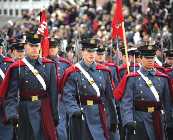 Cadets in uniform march with swords and flags with stadium audience in the background