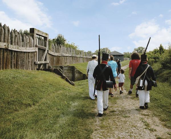 A group of tourists and two War of 1812 reenactors walk along a stone path next two a wooden fence. 