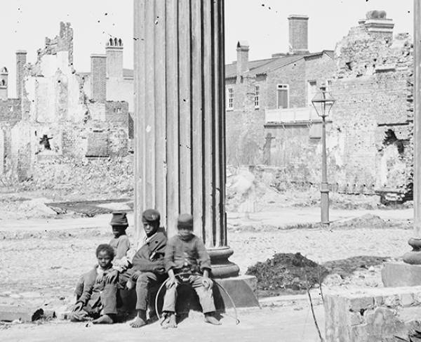 This is a black and white image of four young boys sitting barefoot and dressed in ragged clothing  on the rubble of a battlefield. 