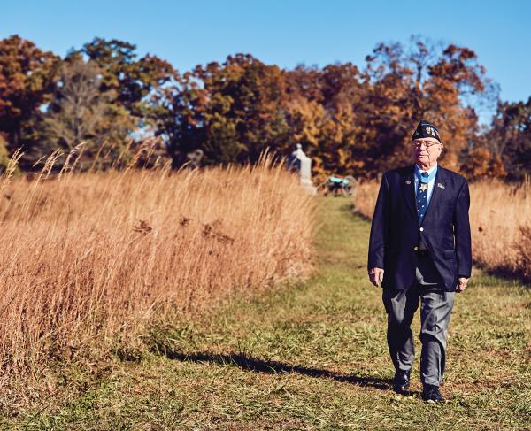 World War II Veteran in a blazer and hat walks a field with a monument in the background