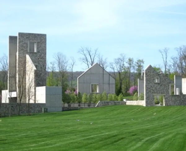 A cluster of modern stone and concrete buildings sits on a gently sloping green lawn. The structures have clean geometric lines, including tall rectangular towers and gabled‑roof sections. 