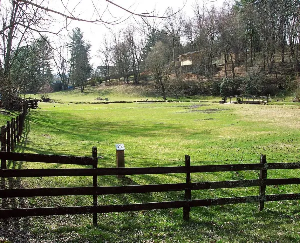 A green field inside a wooden fence with trees and homes visible in the background. Preserved portion of the original muster field where Virginia patriot militia gathered on September 23, 1780.