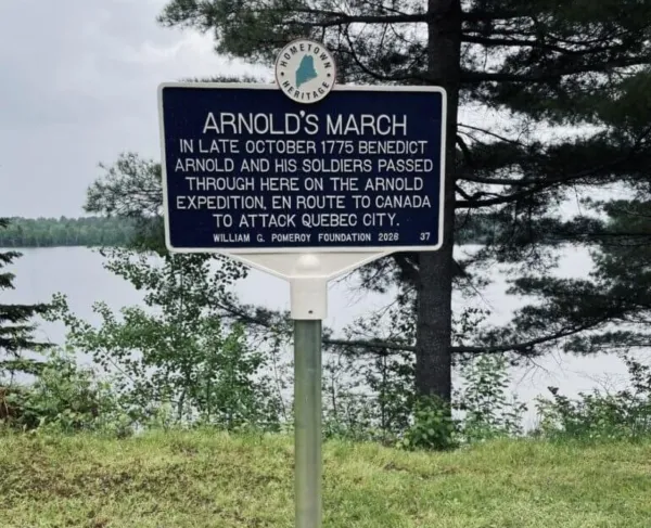 A blue “Arnold’s March” historical marker stands on a grassy lakeside clearing, surrounded by trees under a clear sky.