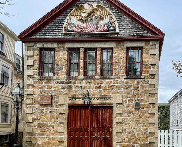 A small historic stone armory built of rough gray blocks with red trim along the gabled roof. Centered near the roof is a large carved and painted patriotic emblem featuring an eagle with outstretched wings and crossed American flags.