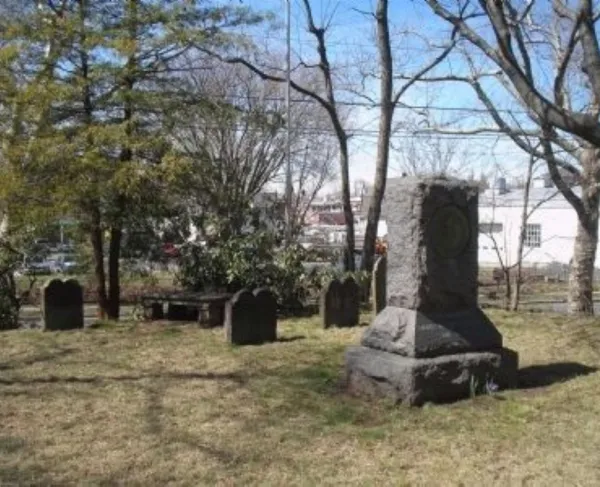 A large stone monument with an engraved circular emblem stands among rows of headstones in a small grassy cemetery bordered by leafless trees and nearby buildings.
