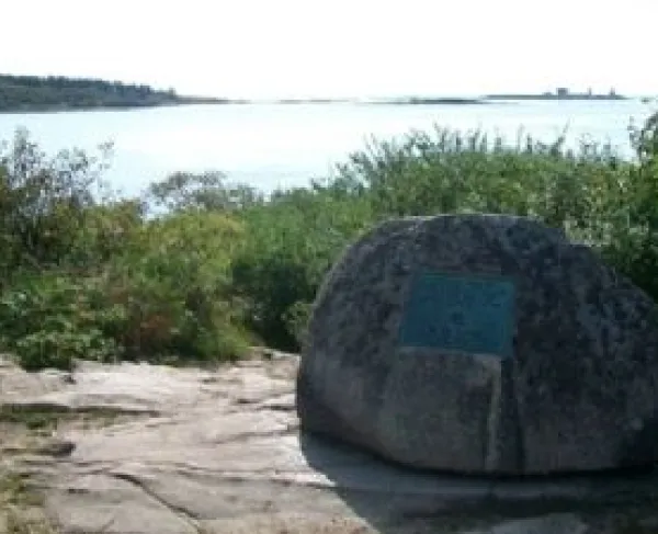 A large rock with a metal plaque sits on a rocky shoreline overlooking calm water, surrounded by low greenery and distant tree‑covered land.