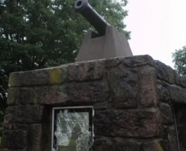 A black cannon sits atop a small stone structure with a glass window, surrounded by green trees under an overcast sky.