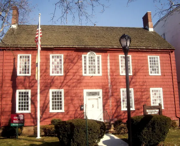 A red historic house with white trim and twin brick chimneys stands behind a flagpole and walkway lined with bushes.