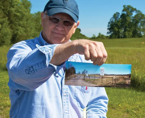 Historian Bud Hall holds a picture of the "for sale" signs and development proposals that once threatened the land where he's standing, now owned by the Trust.