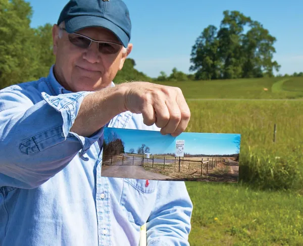 Historian Bud Hall holds a picture of the "for sale" signs and development proposals that once threatened the land where he's standing, now owned by the Trust.