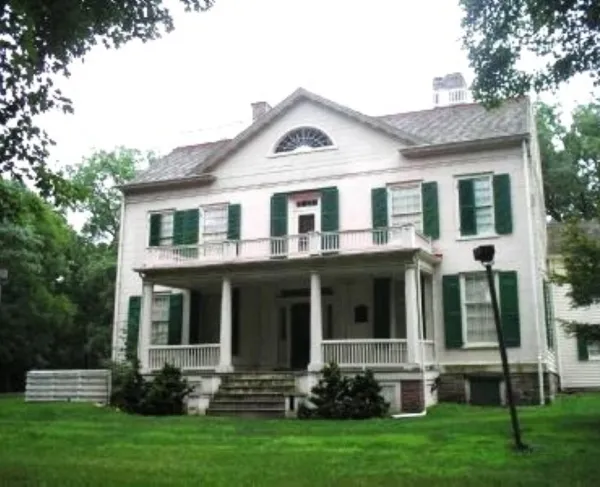 A large white historic house with green shutters, a columned front porch, and a balcony stands on a grassy lawn surrounded by trees.