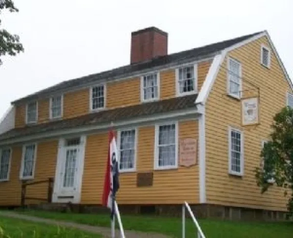 A two‑story yellow wooden house with white trim stands behind a small walkway. The building has a steep roof, a central brick chimney, and evenly spaced white‑framed windows.