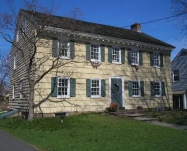 A yellow colonial‑style house with green shutters and a steep dark roof stands behind a leafless tree, with a stone walkway leading to its green front door.