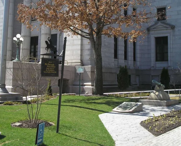 A light‑stone courthouse stands behind a grassy area with a tree, a small cannon monument, and a historical marker noting the site of an early borough courthouse built before the Revolution.