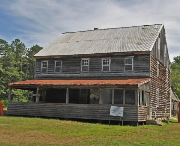 An old two‑story wooden building with a rusted metal porch roof stands in a grassy clearing.