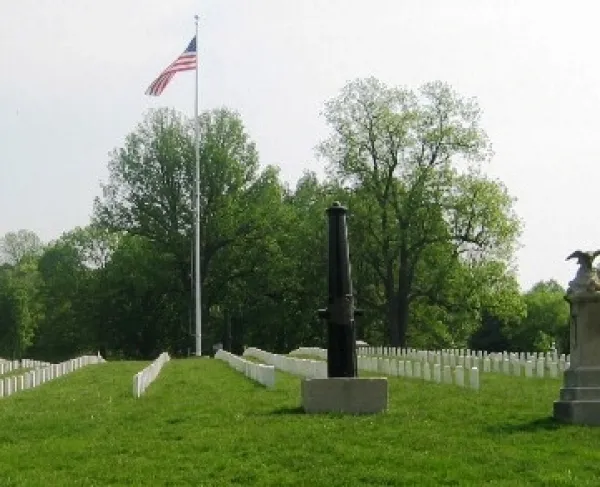 Rows of white headstones line a green cemetery lawn, arranged in straight, even rows. A tall flagpole flying the United States flag stands in the center background. In the foreground, a vertical cannon monument and a stone marker topped with an eagle sculpture sit among the grave.