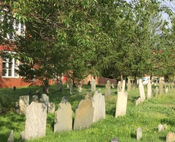 Historic cemetery with rows of weathered gravestones standing in a grassy, tree-shaded burial ground beside a large red-brick building, illuminated by late afternoon sunlight.