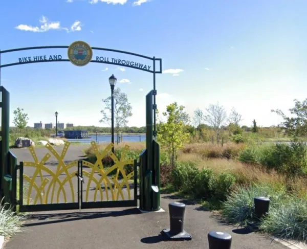 A decorative metal gateway features tall yellow metal shapes resembling stylized reeds, with a circular emblem above of the Seal of Elizabeth.  Beyond the gate, the path leads toward a waterfront lined with trees, shrubs, lampposts, and distant buildings under a bright blue sky.