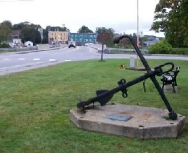 A large black metal anchor sits upright on a square concrete base in a small grassy roadside park. A bronze plaque is mounted on the front of the base, and a wooden bench stands nearby. 