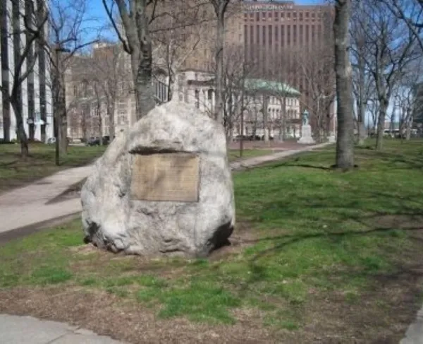A large boulder with a metal plaque stands in a grassy urban park surrounded by trees and tall buildings.